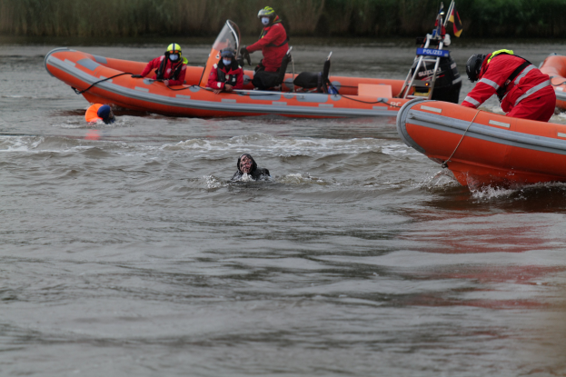Eine Gruppe von Menschen in einem aufblasbaren Boot auf einem Fluss, mit zwei Personen im Wasser im Vordergrund und Vegetation im Hintergrund, alle mit Schwimmwesten und Helmen bei einer Rettungsaktion.