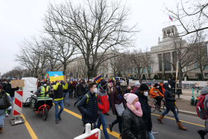 Eine große Gruppe von Menschen nimmt an einer Protestmarsch auf einer Straße in Washington, D.C. teil, einige halten Schilder und Banner, andere fahren Fahrräder, und Schilder, Bäume und ein klarer blauer Himmel sind im Hintergrund zu sehen.