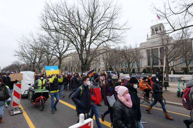 Eine große Gruppe von Menschen nimmt an einer Protestmarsch auf einer Straße in Washington, D.C. teil, einige halten Schilder und Banner, andere fahren Fahrräder, und Schilder, Bäume und ein klarer blauer Himmel sind im Hintergrund zu sehen.
