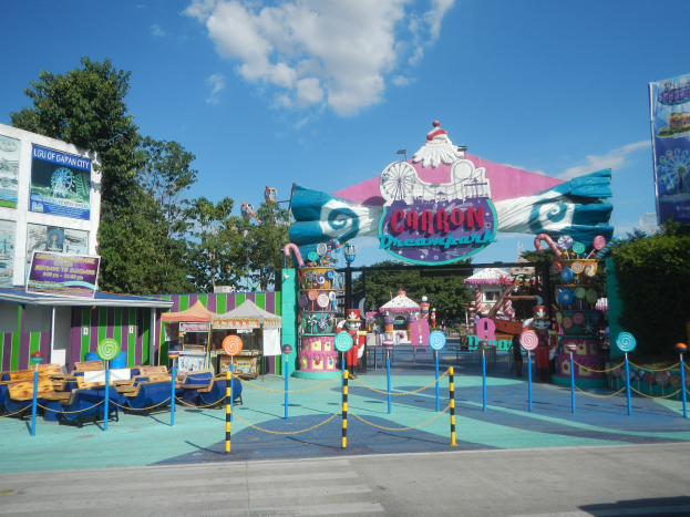 Karussell am Parkeingang mit Straße, eingezäuntem Bereich, Buden, Banner, Bäumen und bewölktem Himmel.