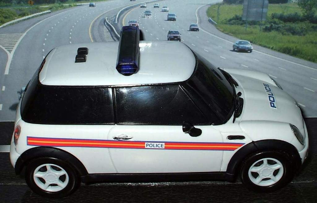 Weißes Polizeiauto mit "Polizei"-Aufschrift auf einer Straße vor einer Stadt, umgeben von fahrendem Verkehr, Gras und Bäumen auf beiden Seiten.