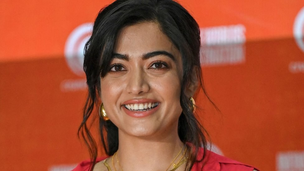 Eine Frau in einem roten Kleid mit einem eleganten Dutt und dezentem Make-up steht strahlend da, trägt schwarze High Heels und ein hellgelbes Banner mit der Aufschrift 'Die besten Schönheits Looks von Sonam Kapoor beim Cannes Film Festival' im Hintergrund.
