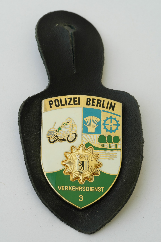 Ein schildförmiges Polizeibadge mit "Polizei Berlin" in fetter schwarzer Schrift auf einem blauen Hintergrund mit einem weißen Stern und Rahmen, das auf einer Oberfläche ruht.