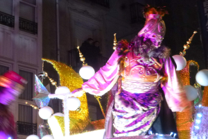 Ein Mann in einem Kostüm steht auf einem geschmückten, beleuchteten Festwagen in einer Stadtparade, im Hintergrund ist ein Gebäude mit Fenstern und Geländern zu sehen.