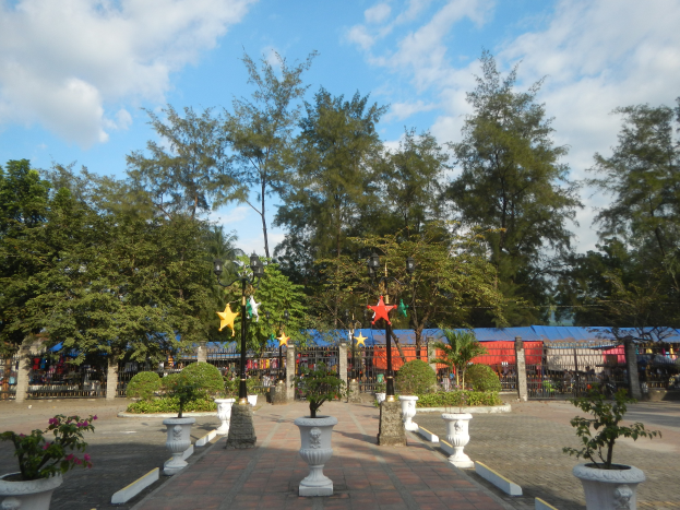 Ein Park mit Topfpflanzen und Bäumen in der Mitte, umgeben von Straßenlaternen, Schmuckelementen und Grills unter einem bewölkten Himmel.