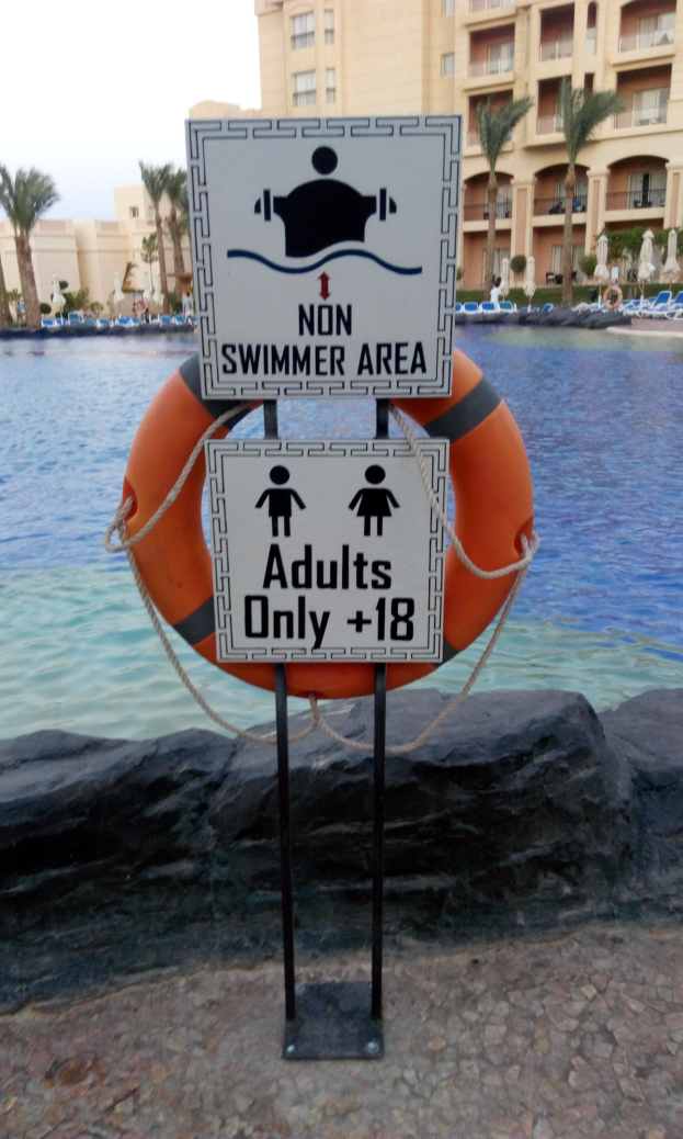 Ein Schwimmbad mit einem Schild "Nur für Erwachsene - Kein Schwimmer" im Vordergrund, umgeben von Bäumen, Gebäuden und einem klaren blauen Himmel, mit einem Rettungsring in der Mitte des Pools.