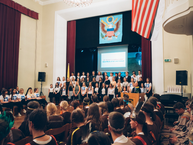 Eine Gruppe von Menschen steht an einem Podium mit einem Mikrofon vor einem sitzenden Publikum während eines 2. Kunst- und Kulturwochen-Events, mit Fahnen, Lautsprechern, einem Bildschirm mit Text und einem Bild sowie Vorhängen und einem Kronleuchter im Hintergrund.