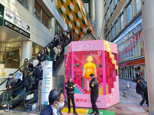 Eine Gruppe von Menschen steht vor einem gläsernen Gebäude mit Säulen und Texttafeln, einige tragen Masken, während eine Rolltreppe mit Menschen im Hintergrund zu sehen ist; ein pinker Kasten in der Mitte zeigt Schaufensterpuppen und trägt die Aufschrift "Netflix's new Netflix booth in Tokyo."