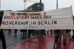 Eine Gruppe von Menschen steht auf der Straße und hält ein Protestplakat in der Hand, auf dem "Die Wrangellstraße 5 protestiert gegen den Ausverkauf in Berlin" steht, mit Fahrrädern, Pfählen, Gebäuden und einem Kran im Hintergrund und einem sichtbaren Himmel.