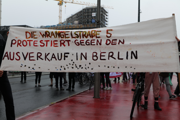Eine Gruppe von Menschen steht auf der Straße und hält ein Protestplakat in der Hand, auf dem "Die Wrangellstraße 5 protestiert gegen den Ausverkauf in Berlin" steht, mit Fahrrädern, Pfählen, Gebäuden und einem Kran im Hintergrund und einem sichtbaren Himmel.