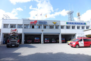 Feuerwache mit mehreren Feuerwehrautos vor dem Gebäude, Gebäude mit Fenstern und Text, Flaggen mit Stangen im Hintergrund, bewölkter Himmel und fernes Turm.