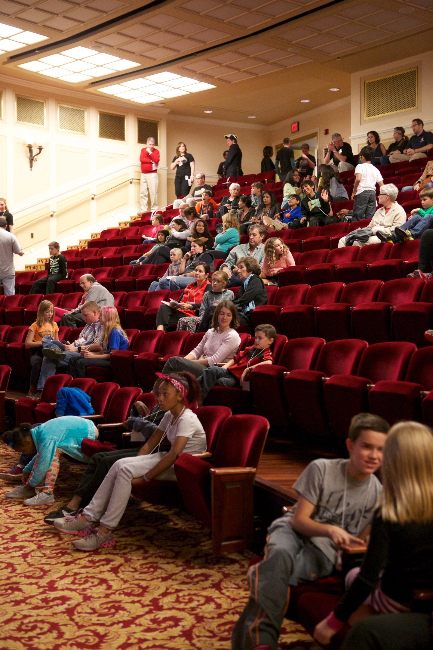 Eine große Gruppe von Menschen, darunter Kinder, die auf Stühlen sitzen und stehen, umgeben von verstreuten Stühlen, Wandlampen und Deckenlampen.