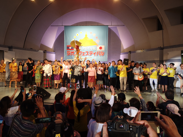 Gruppe von Menschen auf einer Bühne mit Kameras, Handys und Musikinstrumenten während einer Festival-Eröffnung, mit einem Banner, Wänden und Deckenlampen im Hintergrund.