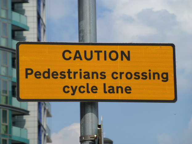 Gelbes Warnschild an einem Pfahl vor einem Gebäude, auf dem steht "Fußgänger überqueren Radweg" vor einem klaren blauen Himmel.