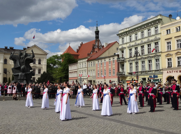 Eine Gruppe von Menschen in weißen Kleidern steht vor einer Menge, einige halten Musikinstrumente, mit Gebäuden, Bäumen, einer Statue, Fahnen und einem bewölkten Himmel im Hintergrund, wahrscheinlich während des Nationalfeiertags der Tschechischen Republik.