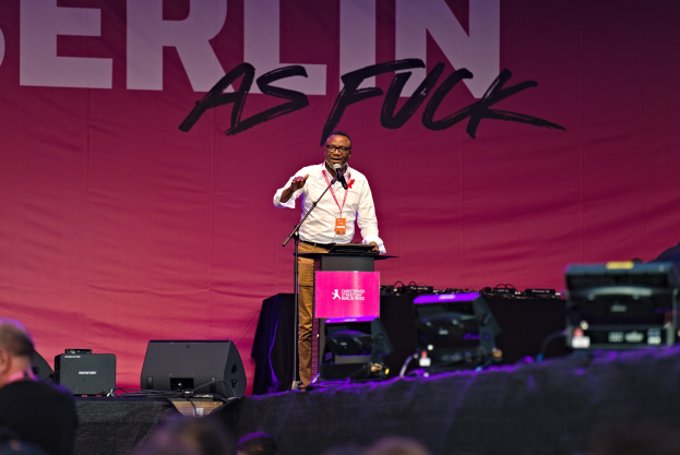 Ein Mann in einem weißen Hemd und braunen Hosen steht auf einer Bühne und hält ein Mikrofon in der Hand, mit einem pinken Banner mit der Aufschrift "Berlin als Fuck" dahinter und einer Menge davor.