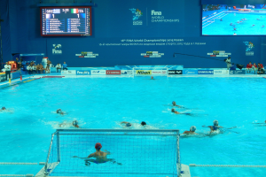 Eine Gruppe von Menschen spielt Wasserball in einem große Schwimmbecken mit einem Netz in der Mitte, Zuschauern auf der linken Seite und einer Wand mit einem Bildschirm mit Text und Logos im Hintergrund bei den FINA-Weltmeisterschaften 2016 in Kazan.