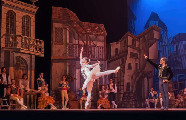 Zwei Tänzer performen auf einer Bühne im Royal Opera House in London, mit einem Publikum im Vordergrund und einem Bildschirm mit Gebäudebildern im Hintergrund.