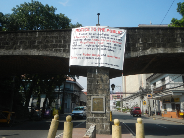 Öffentlicher Hinweisschild an der Seite einer Brücke über eine Straße mit Fahrzeugen, mit Gebäuden, Bäumen, Strommasten und Drähten im Hintergrund unter einem klaren blauen Himmel.