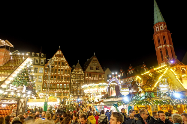 Ein geschäftiger Weihnachtsmarkt in Nürnberg, Deutschland mit Menschen, die sich um geschmückte Stände versammeln, Gebäuden mit Fenstern und einem Uhrenturm im Hintergrund und einem dunklen Himmel.