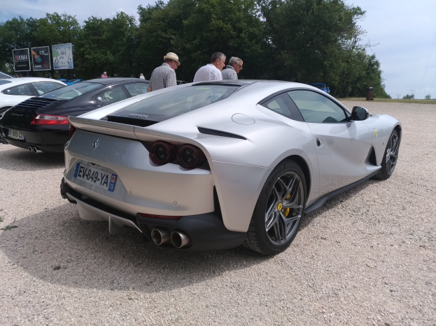 Roter Ferrari 812 Superfast auf dem Goodwood Festival of Speed, umgeben von Menschen mit Mützen, Bäumen, Schildern und einem bewölkten Himmel im Hintergrund.