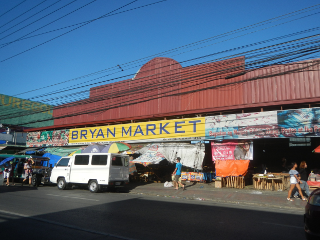 Ein geschäftiger Marktplatz mit Fahrzeugen, Fußgängern, Marktständen, Bannern, Schirmen und hölzernen Gegenständen, mit einem "Bryan Market"-Schild im Hintergrund unter einem Himmel mit Stromleitungen.