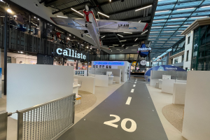 Ein großer Raum mit weißen Wänden, ein vom Himmel hängendes Flugzeug, ein Geländer auf der linken Seite, einige Gegenstände auf der rechten Seite, Texttafeln im Hintergrund und an Metallstangen an der Decke befestigte Lampen, wahrscheinlich am Düsseldorf Airport.