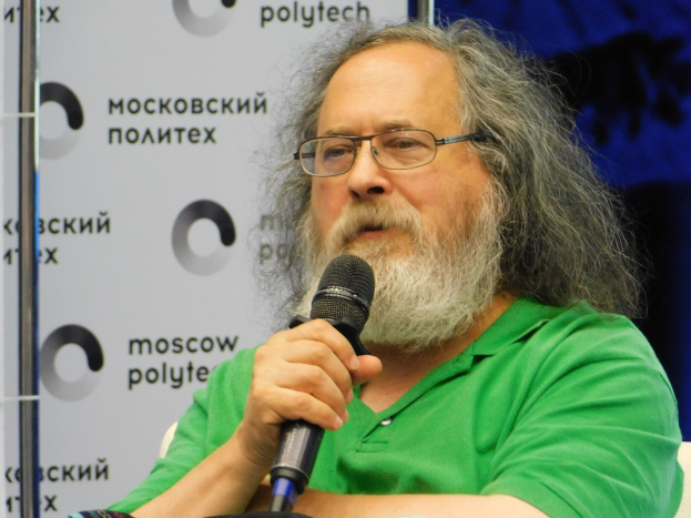 Ein Mann mit langen Haaren und Brille trägt ein grünes T-Shirt und sitzt auf einem Stuhl, auf dem er ein Mikrofon hält. Im Hintergrund ist ein Banner zu sehen, das ihn als Psycholinguisten ausweist.