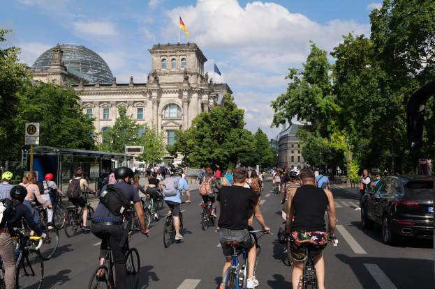 Eine Gruppe von Menschen, die auf Fahrrädern eine von Bäumen gesäumte Straße in Berlin, Deutschland, entlangfahren, mit Gebäuden und einer Bushaltestelle, unter einem bewölkten Himmel und einer Flagge, die auf einem Gebäude weht.