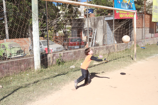 Ein junger Junge schießt einen Fußball in ein Tor, mit einem Netz, Pfosten, Bannern, einer Wand, Fahrzeugen, Bäumen, Gebäuden und einem klaren blauen Himmel im Hintergrund.