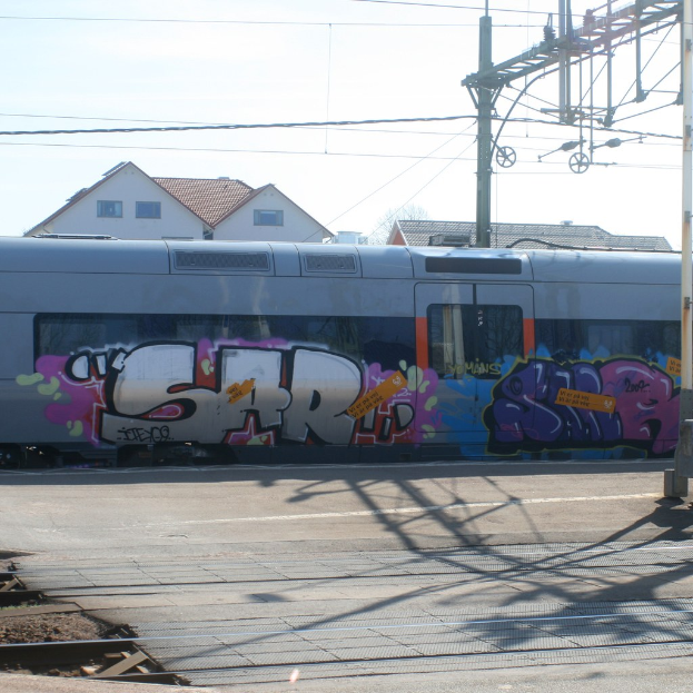 Graffiti an einem Zug mit sichtbaren Kabeln und Häusern mit Fenstern im Hintergrund.
