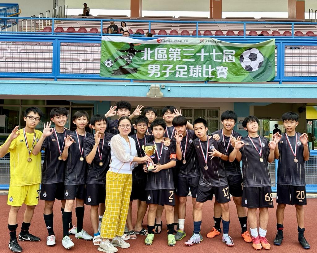 Gruppe junger Männer in Fußballuniformen auf einem Feld stehend, Medaillen tragend und einen Pokal haltend, mit einem Banner im Hintergrund, das 'Yokohama U-16 Boys Soccer Team' lautet, und sitzende Zuschauer in der Nähe.