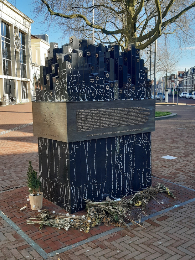 Denkmal in einem Stadtplatz gewidmet an Holocaust-Opfer, mit einem Baum davor, einer Topfpflanze, verstreuten trockenen Blättern und Gebäuden, Fahrzeugen und Bäumen im Hintergrund.