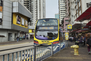 Ein gelber Doppeldeckerbus fährt eine Straße mit hohen Gebäuden entlang, mit Menschen auf dem rechten Gehweg, einem Hydranten und einem Geländer, unter einem klaren blauen Himmel.
