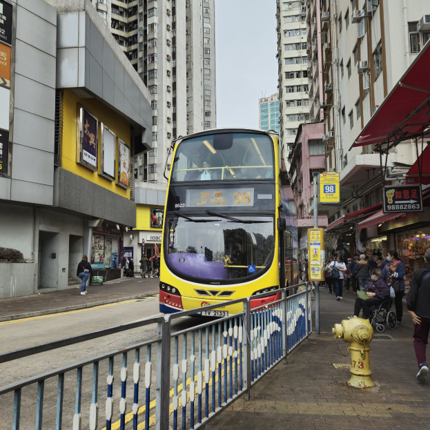 Ein gelber Doppeldeckerbus fährt eine Straße mit hohen Gebäuden entlang, mit Menschen auf dem rechten Gehweg, einem Hydranten und einem Geländer, unter einem klaren blauen Himmel.
