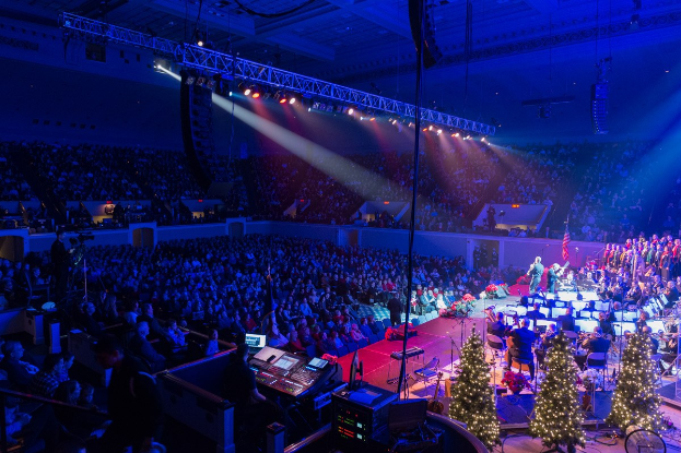 Eine große Menschenmenge in einem geschmückten Auditorium schaut einer Band zu, die auf einer Bühne mit Weihnachtsbäumen und Lichterketten spielt.