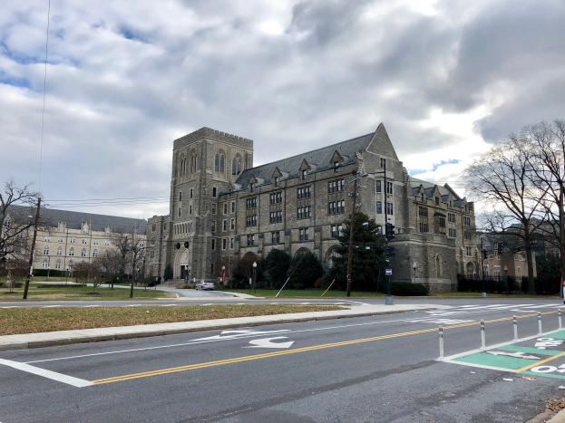 Großes Gebäude mit zahlreichen Fenstern, umgeben von Bäumen, Laternen, Ampeln und Fahrzeugen auf der Straße, unter einem bewölkten Himmel, als der neue Campus der Universität Pennsylvania identifiziert.