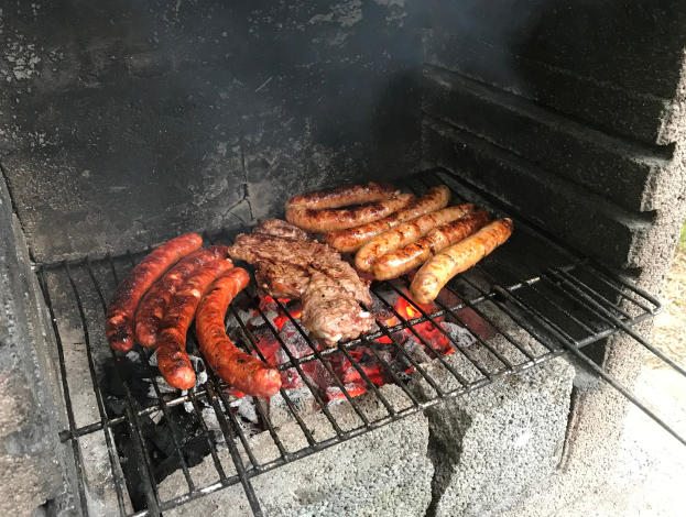 Grill mit Würstchen und Fleisch darauf, umgeben von brennender Kohle und Flammen, draußen auf Gras stehend.