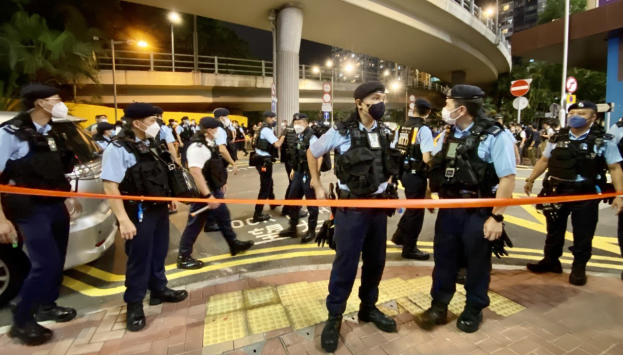 Polizeibeamte in Uniform und Mützen stehen vor einer Menge, mit einem Band im Vordergrund und Fahrzeugen links, in einer Szene, die wahrscheinlich auf eine Hongkonger Protest follows, bei der ein Mann erschossen und getötet wurde.