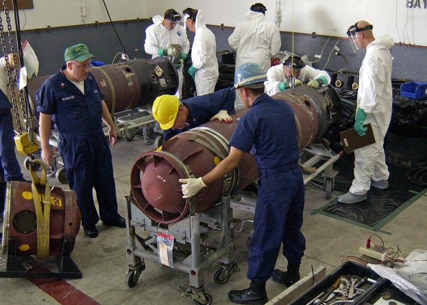 Männer in Helmen und Handschuhen bei der Arbeit an einer großen Bombe in einer Fabrik mit Maschinen und Wandtext.
