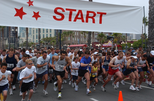 Gruppe von Läufern bei einem Marathon, die an einer Verkehrskegel vorbeilaufen, mit einem Banner und Gebäuden im Hintergrund bei klarem blauem Himmel.
