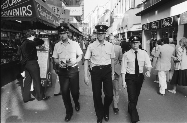 Drei Polizisten in Uniform gehen eine belebte Stadtstraße entlang, die von hohen Gebäuden und Geschäften gesäumt ist, mit Passanten auf beiden Seiten.