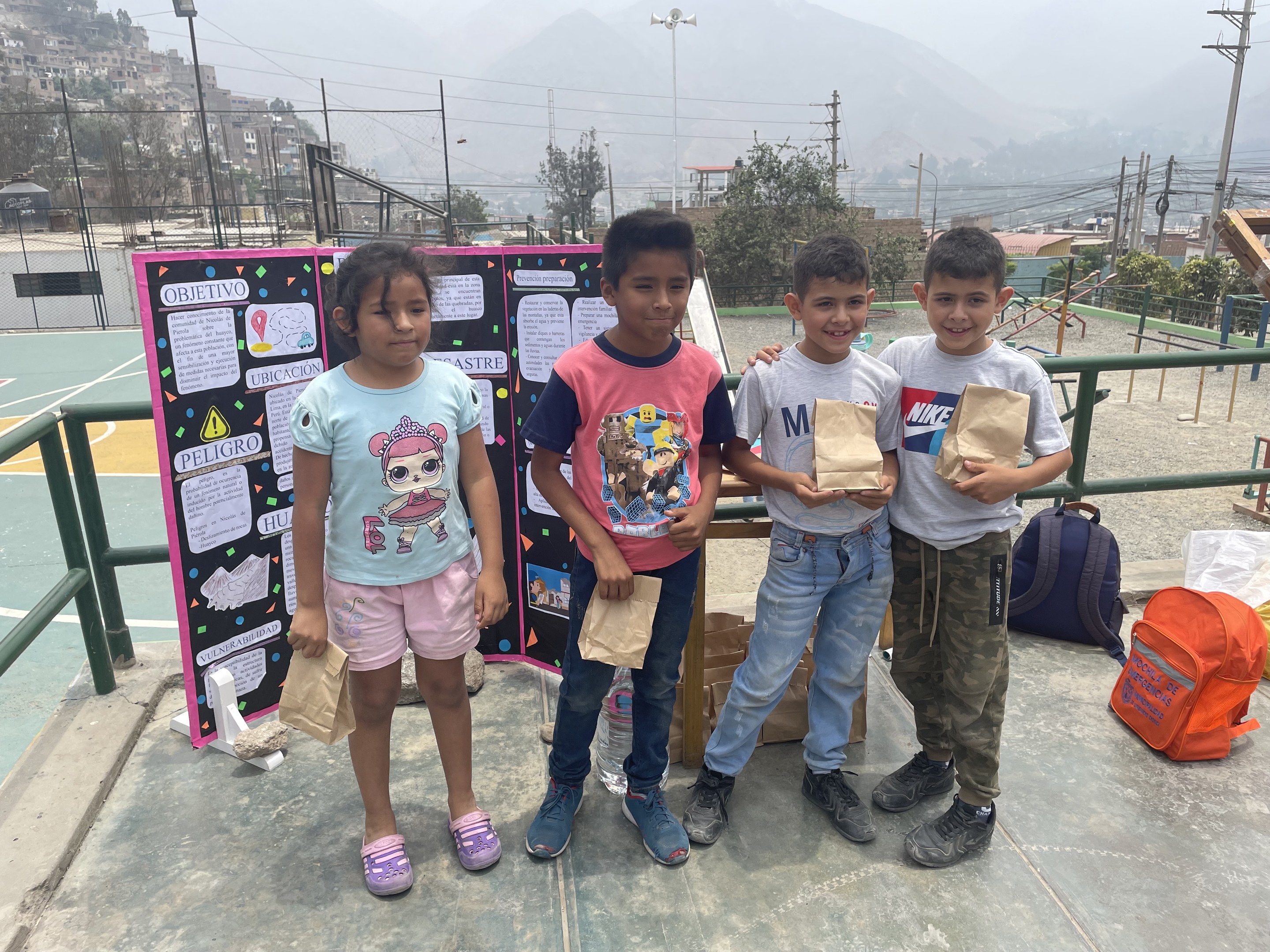 Gruppe von Kindern auf einem Basketballfeld mit Papiertüten in den Händen, mit Vorräten auf dem Boden und einer Tafel mit Text links daneben, vor einem Hintergrund aus Zäunen, Pfosten, Straßenlaternen, Bäumen, Gebäuden, Hügeln und Himmel.