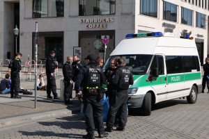 Eine Gruppe von Polizeibeamten steht vor einem Starbucks-Café, rechts parkt ein Van, links stehen und sitzen Menschen, im Hintergrund ein Gebäude mit Fenstern, ein Schild, eine Laterne und ein Zaun.