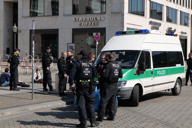 Eine Gruppe von Polizeibeamten steht vor einem Starbucks-Café, rechts parkt ein Van, links stehen und sitzen Menschen, im Hintergrund ein Gebäude mit Fenstern, ein Schild, eine Laterne und ein Zaun.