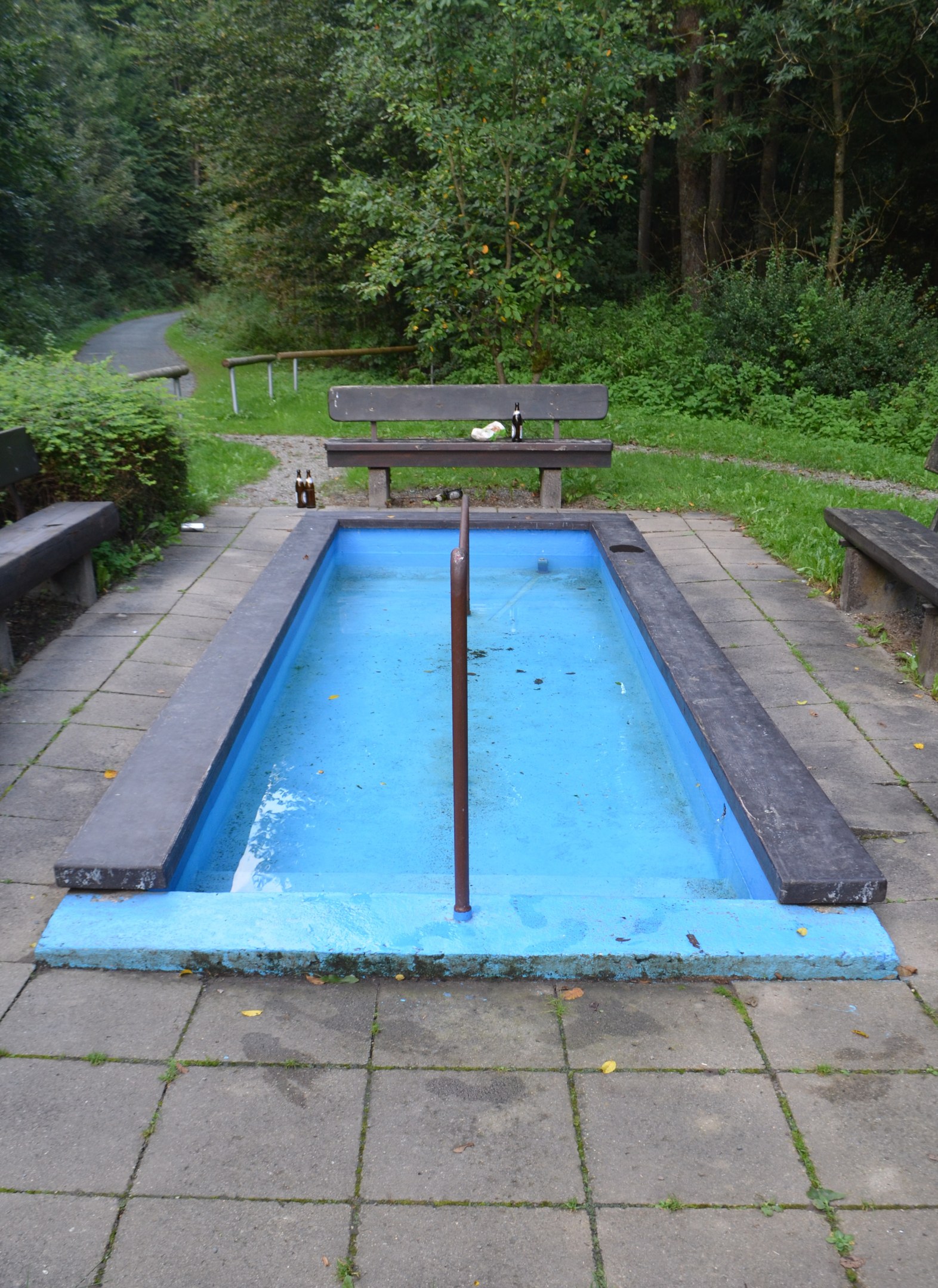 Kleiner Parkteich mit zentralem Pfahl, umgeben von B├Ąnken, Grün, Stra├če und B├Ąumen.