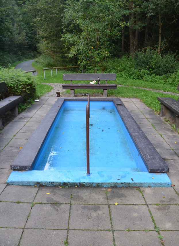 Kleiner Parkteich mit zentralem Pfahl, umgeben von B├Ąnken, Grün, Stra├če und B├Ąumen.