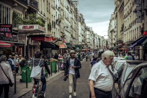 Zwei große Gebäude stehen sich gegenüber auf einer belebten Straße mit Fußgängern, Verkäufern und geparkten Autos, während ein Mann ein Fahrrad fährt und Mülltonnen in der Nähe der Gebäude stehen.