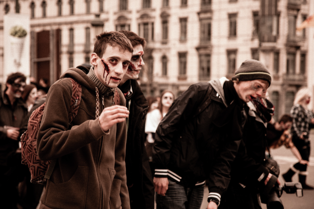 Eine Gruppe von Menschen in Zombie-Kostümen geht eine Straße entlang, mit einem Gebäude im Hintergrund und einer Person mit einer Kamera.