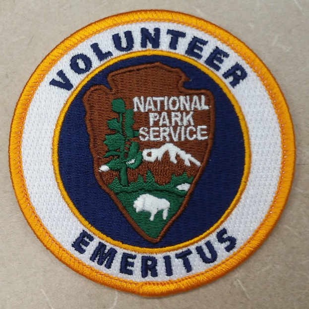 Runder Aufnäher mit blauem Rand und weißem Hintergrund, darauf in der Mitte in schwarzer Schrift 'Volunteer National Park Service Emeritus' in einem gelben Kreis.